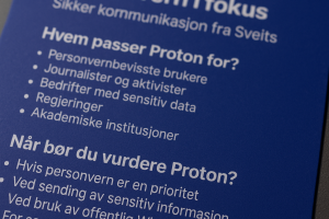 Et nærbilde av en fysisk Proton-plakat med fokus på personvern, fotografert i skrå vinkel med profesjonell belysning, tydelig logo og punkterte lister over hvem Proton passer for og når det bør vurderes.